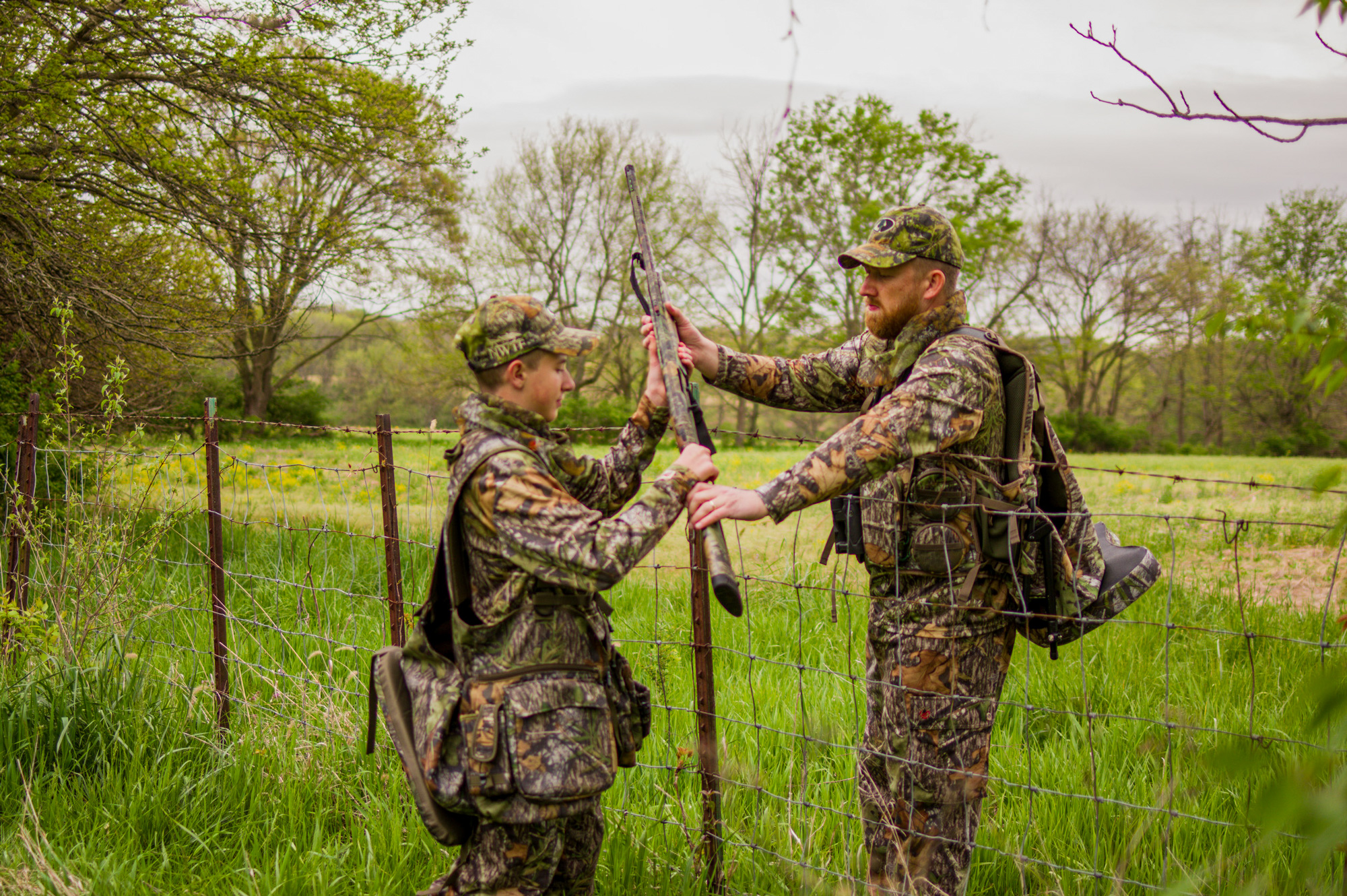 adult hunter handing a gun across a fence to a youth hunter