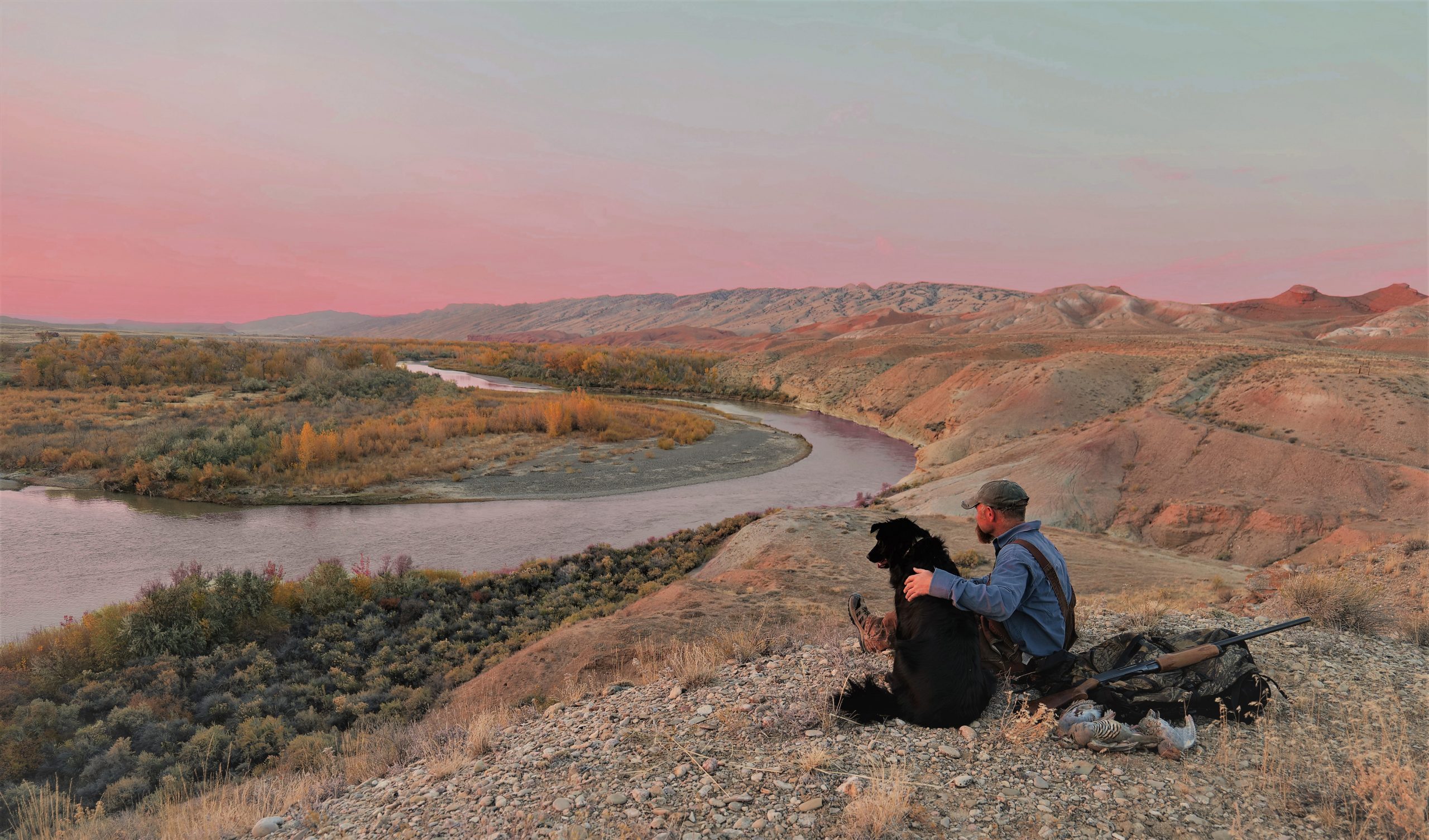 A hunter stares off with his hunting dog at a waterway near a grassland